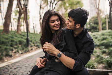 A man embraces a woman from behind in a park, as they gaze lovingly at each other.